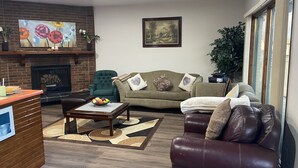 A living room with a fireplace, a green armchair, a brown leather sofa, and a coffee table with fruit. - Ranchland Inn (Nanton)