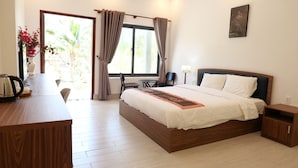 Ein Hotelzimmer mit einem hölzernen Bett, einem Schreibtisch mit Wasserkocher, einem Sessel, einem kleinen Tisch und Blick auf Grünfläche.