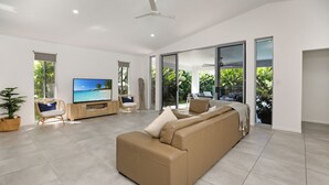 A modern living room with a large sofa, a television, and a view of a tropical landscape through large sliding glass doors. - Heated pool Seaside at Palm cove (Cairns)