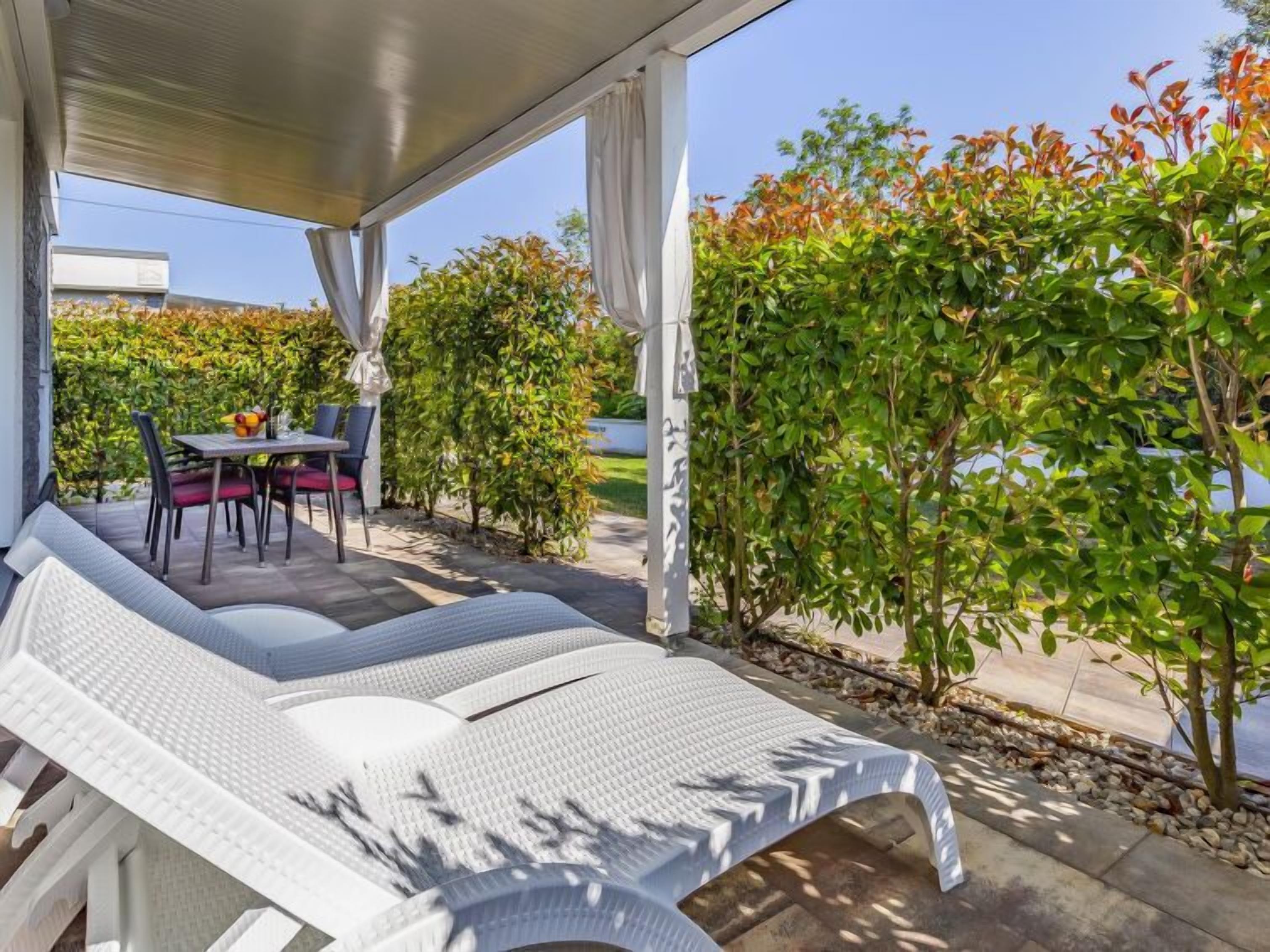 A covered outdoor seating area with a table and chairs, surrounded by greenery and a view of a road.