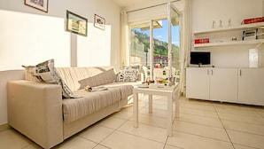 A modern living room with a sofa, a coffee table, and a TV. There are framed pictures on the wall, a window with a view of greenery, and a white cabinet with shelves.