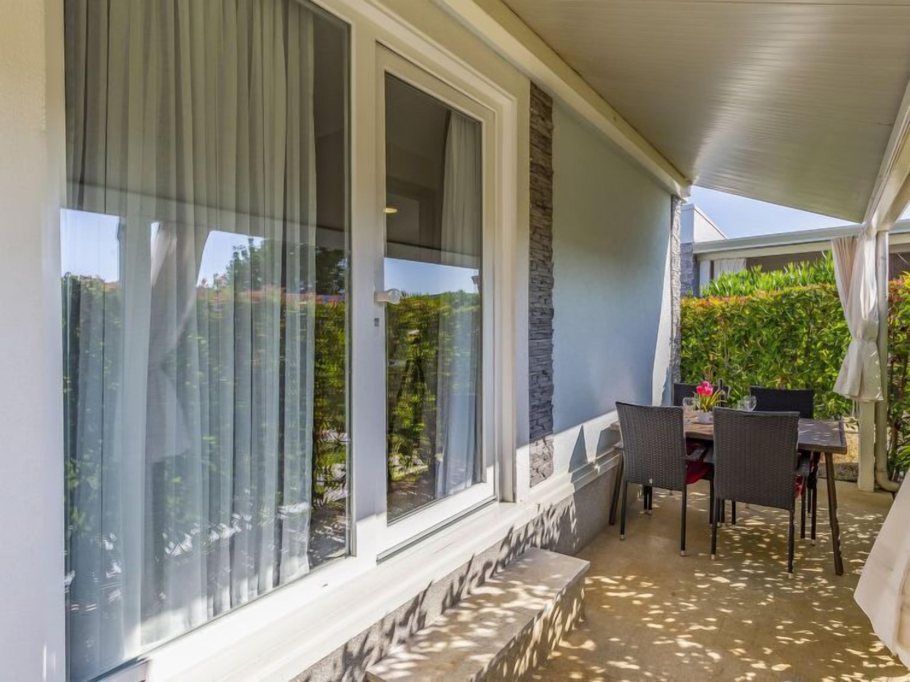 Eine überdachte Terrasse mit Tisch und Stühlen, große Fenster mit Vorhängen und Blick ins Grüne.