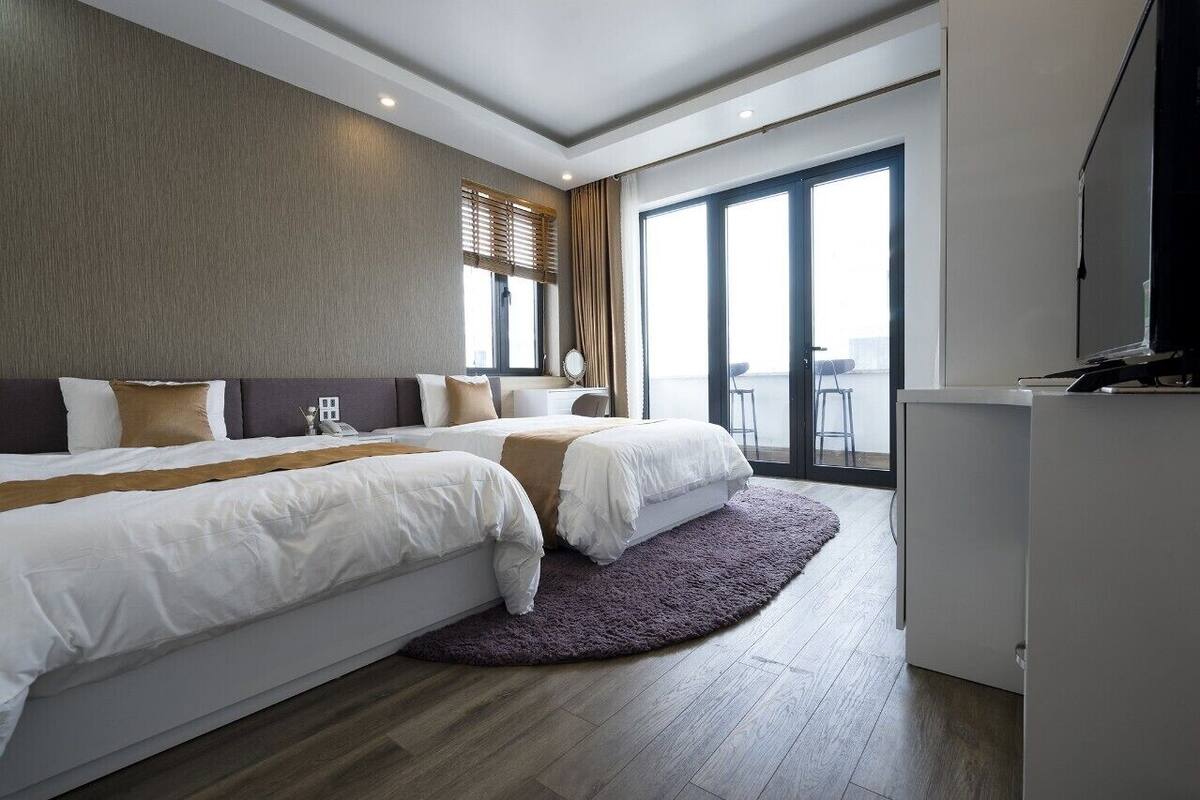 A modern hotel room with two beds, a large window, a balcony, and a flat-screen TV.