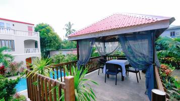 A covered outdoor seating area with a pool, surrounded by lush greenery and a white building with balconies.