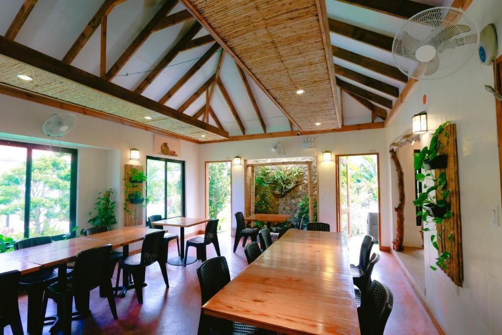 A spacious room with a high ceiling, wooden beams, and large windows. There are multiple tables and chairs arranged for dining or meetings.