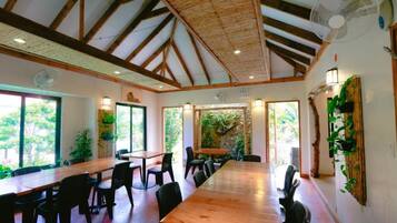 A spacious room with a high ceiling, wooden beams, and large windows. There are multiple tables and chairs arranged for dining or meetings.