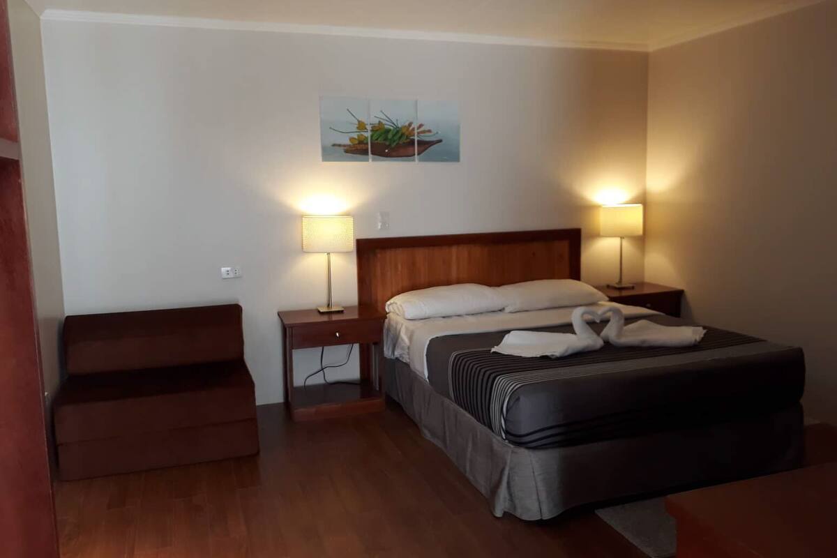 Habitación de hotel con cama, dos mesitas de noche con lámparas, cabecero de madera, un pequeño banco y un cuadro colgado en la pared.