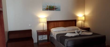 A hotel room with a bed, two bedside tables with lamps, a wooden headboard, a small bench, and a wall-mounted painting.