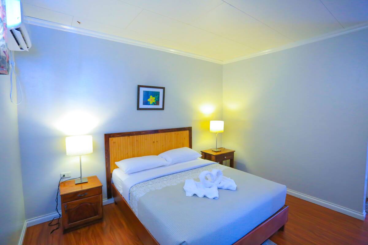 Una cama bien tendida con sábanas blancas, cabecera de madera y un par de toallas sobre la cama. Hay una mesita de noche con una lámpara y un cuadro en la pared.