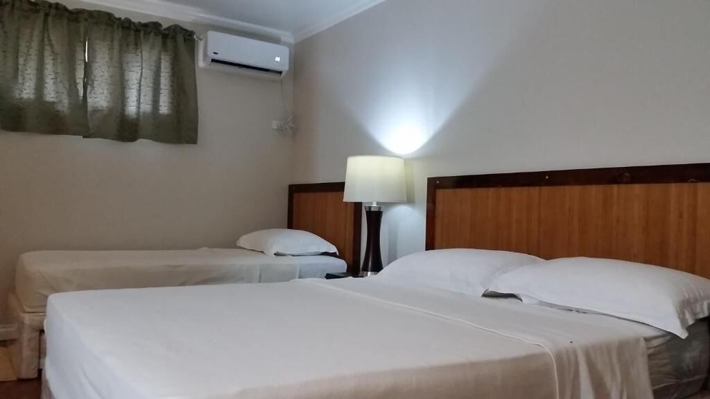 A hotel room with two beds, a nightstand with a lamp, an air conditioning unit, and a window with curtains.