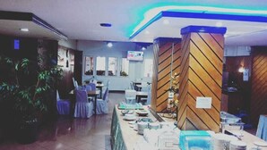 A restaurant with a long counter, tables, chairs, and a television screen. - Rauda Hotel Syariah (Pekanbaru)