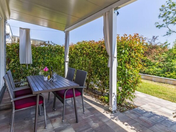 An outdoor patio with a table and chairs, surrounded by greenery and a covered area.