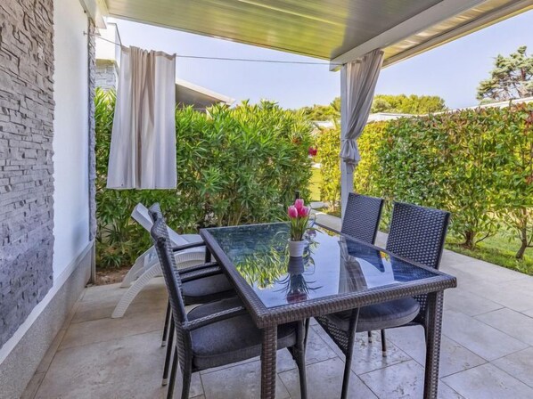 Um pátio externo com mesa de vidro e cadeiras de vime, rodeado por vegetação e uma parede de pedra.