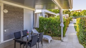 En udendørs patio med et bord og stole, et overdækket siddeområde samt en sti kantet med grønt.