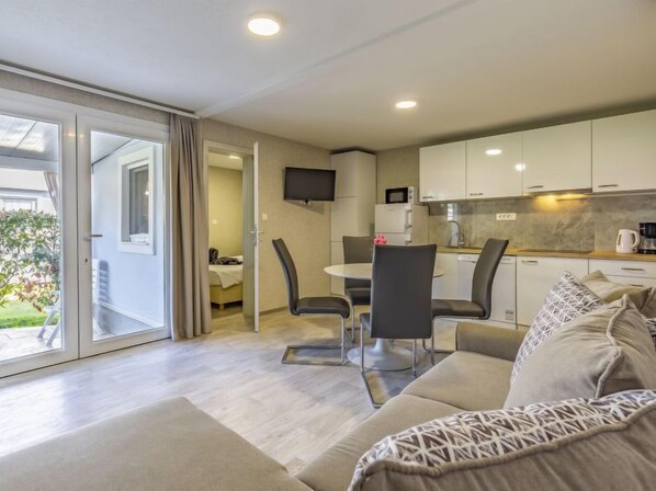 A modern, well-lit room with a sofa, a dining table with chairs, a kitchen area with cabinets and a microwave, and a sliding glass door leading to an outdoor area.
