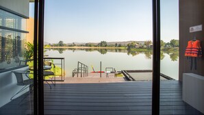 Une vue depuis un balcon doté d’une terrasse en bois donnant sur un lac paisible, avec une chaise et un gilet de sauvetage accroché au mur.