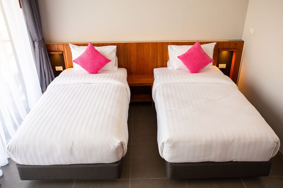 Two single beds with wooden headboards and white bedding, a small wooden nightstand, and a power outlet on the wall.