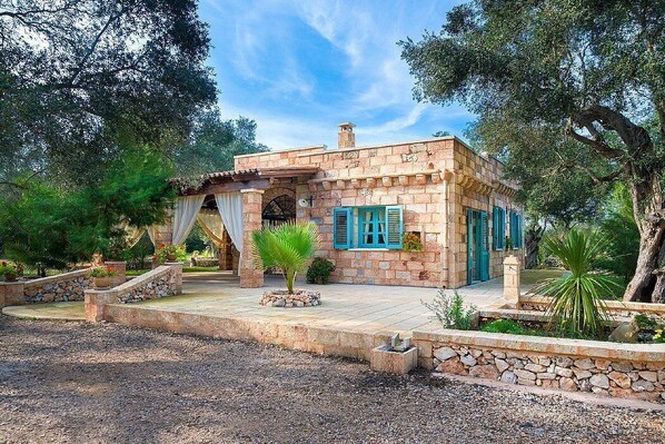 Une maison en pierre avec des volets bleus, entourée d’arbres et bordée d’une allée gravillonnée.