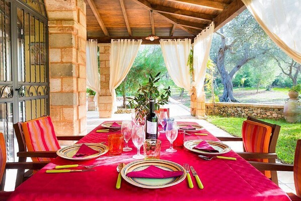 A dining table set outdoors with a red tablecloth, surrounded by stone walls and a view of trees.