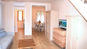 A living room with a sofa, a wooden coffee table, and a TV on a dresser. There is a dining area with a table and chairs, and a staircase leading to another level.