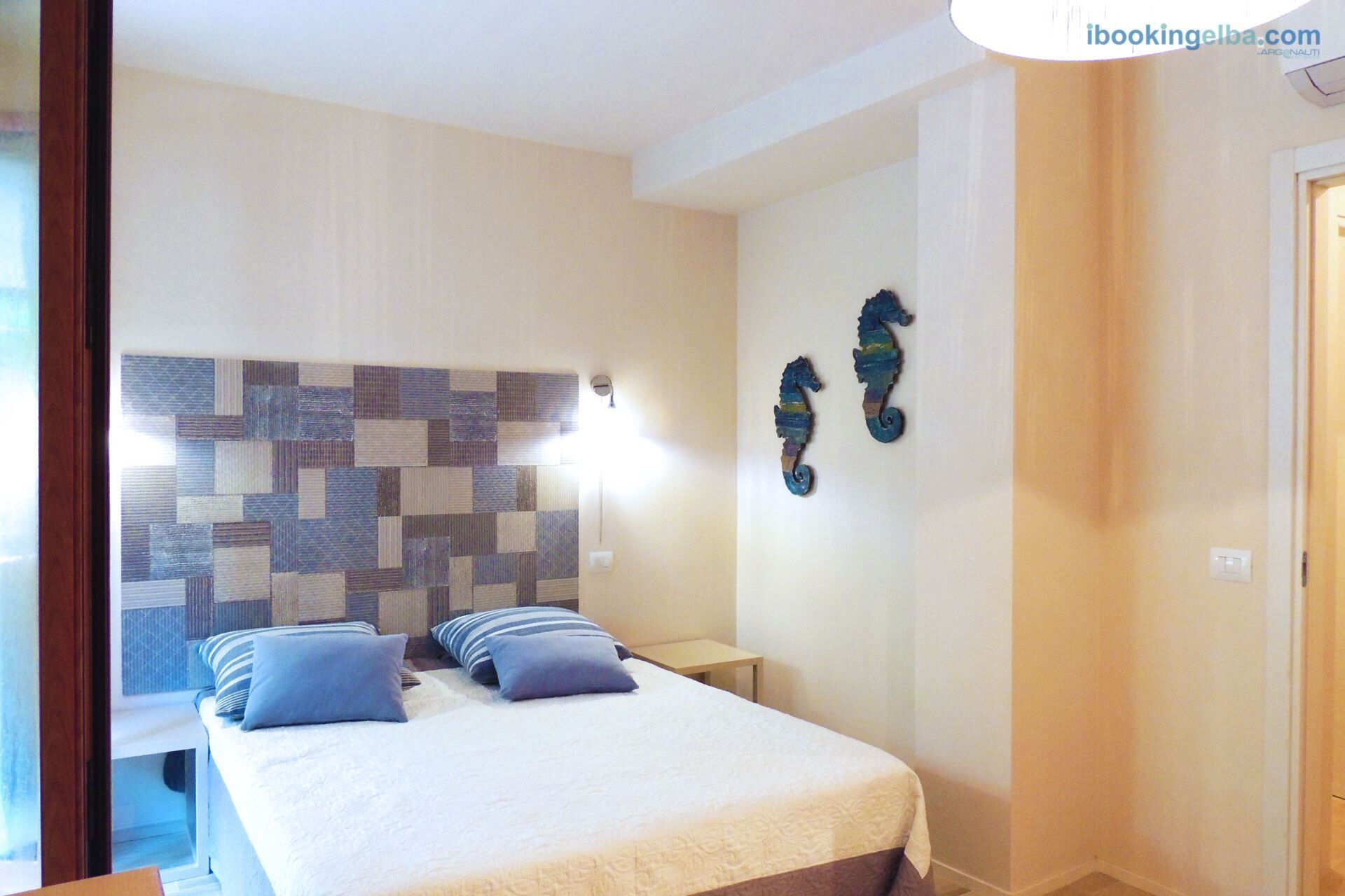 A bedroom with a bed, two pillows, a wall-mounted light, and two decorative wall hangings.