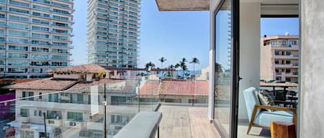 Un balcón con una banca, una silla y vistas a edificios altos y palmeras.
