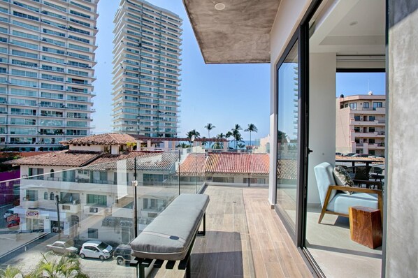 A balcony with a bench, a chair, and a view of tall buildings and palm trees.