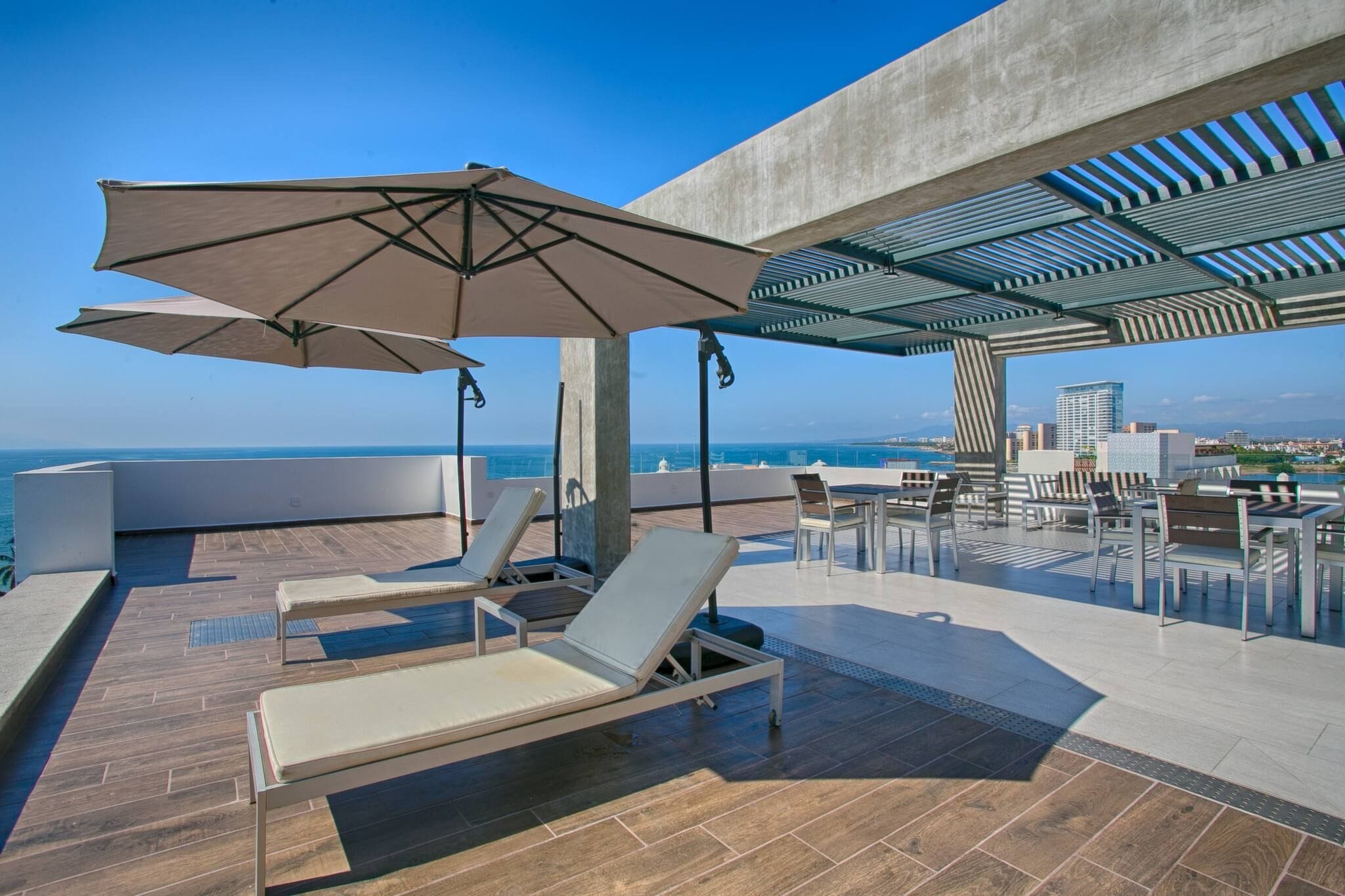 Una terraza en la azotea con sillones de descanso, un paraguas grande y vistas al océano y al horizonte de la ciudad.
