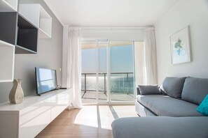 Uma sala de estar moderna com sofá, TV de tela plana e uma grande porta de vidro deslizante que dá para uma varanda com vista para o mar.