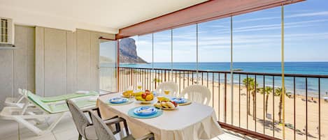 A balcony with a table set for two, overlooking a beach with palm trees and a rocky cliff.