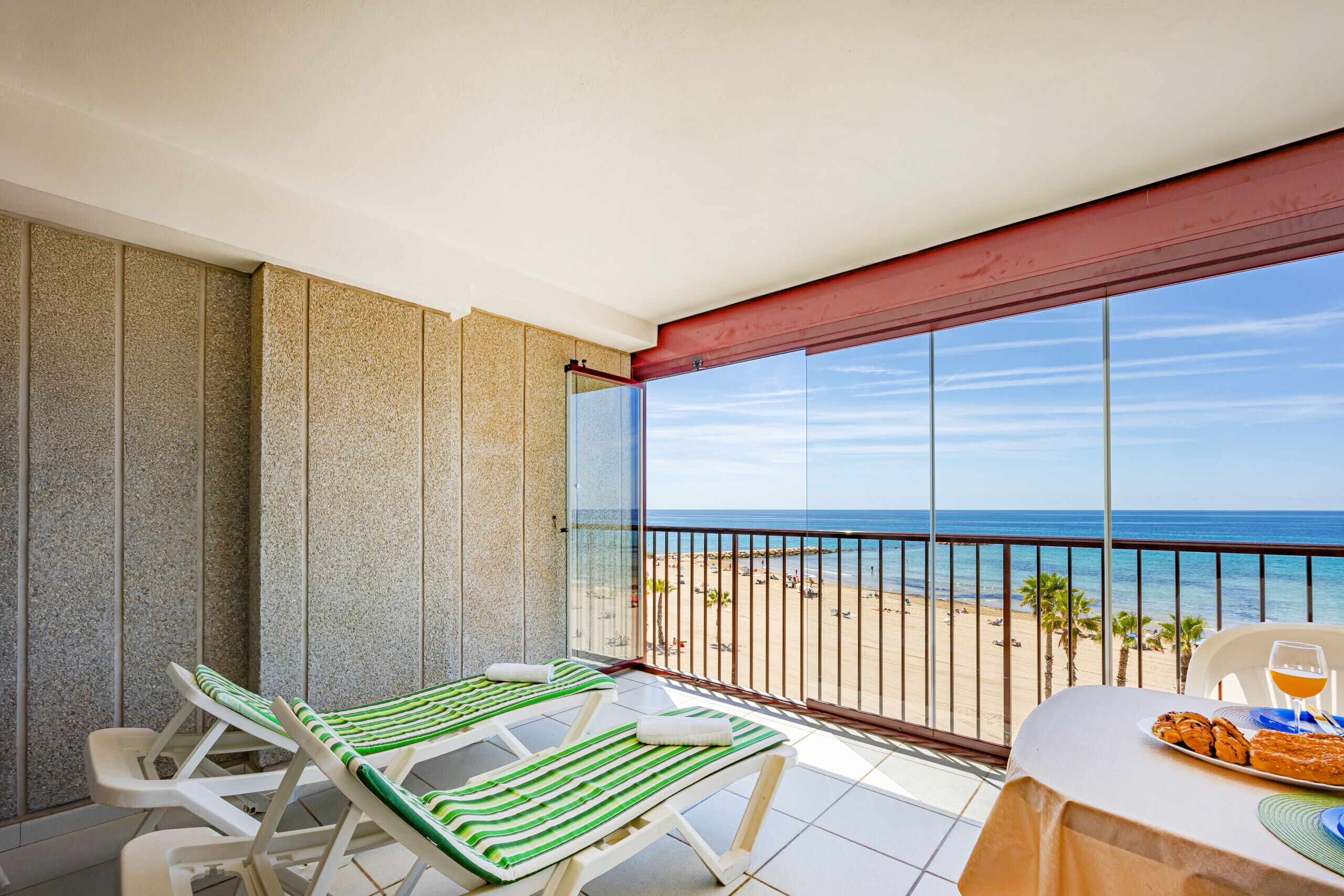 A balcony with two striped lounge chairs, a table with a plate of food and a glass of orange juice, and a view of the beach and ocean.