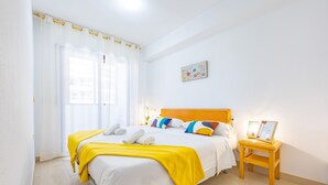 Ein ordentlich bezogenes Bett mit einer gelben Decke, ein Nachttisch aus Holz mit einer Lampe und ein an der Wand hängendes Bild.