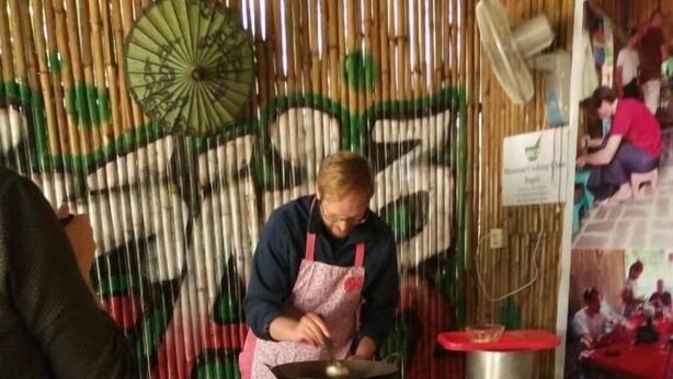 A person cooking on a stove in a rustic kitchen with bamboo walls and a fan. There are blue plastic chairs and a woven basket on the floor. - My Family - Hostel (Nyaung-U)