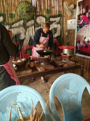 A person cooking on a stove in a rustic kitchen with bamboo walls and a fan. There are blue plastic chairs and a woven basket on the floor. - My Family - Hostel (Nyaung-U)