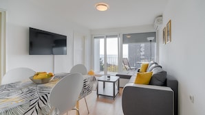 A modern living room with a flat-screen TV, a dining table with chairs, a sofa with cushions, and a view of the outdoors through large windows.