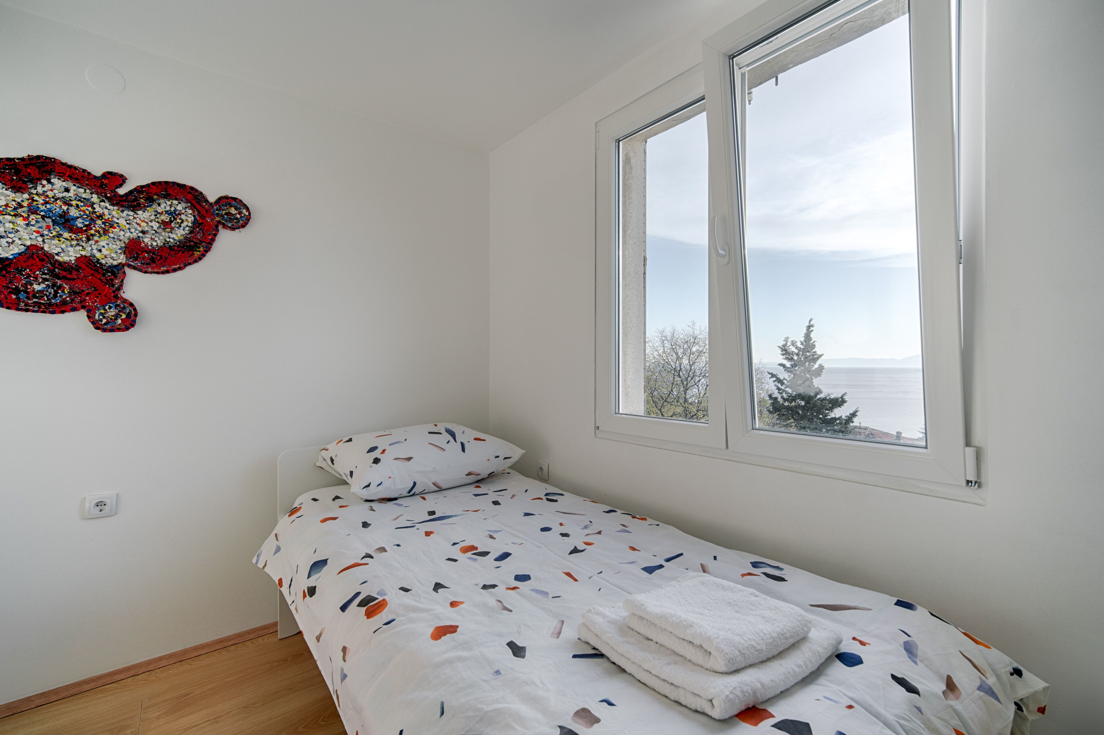 Un lit simple avec une couvre-lit à motifs, une fenêtre donnant sur les arbres et le ciel, et une œuvre d’art accrochée au mur.