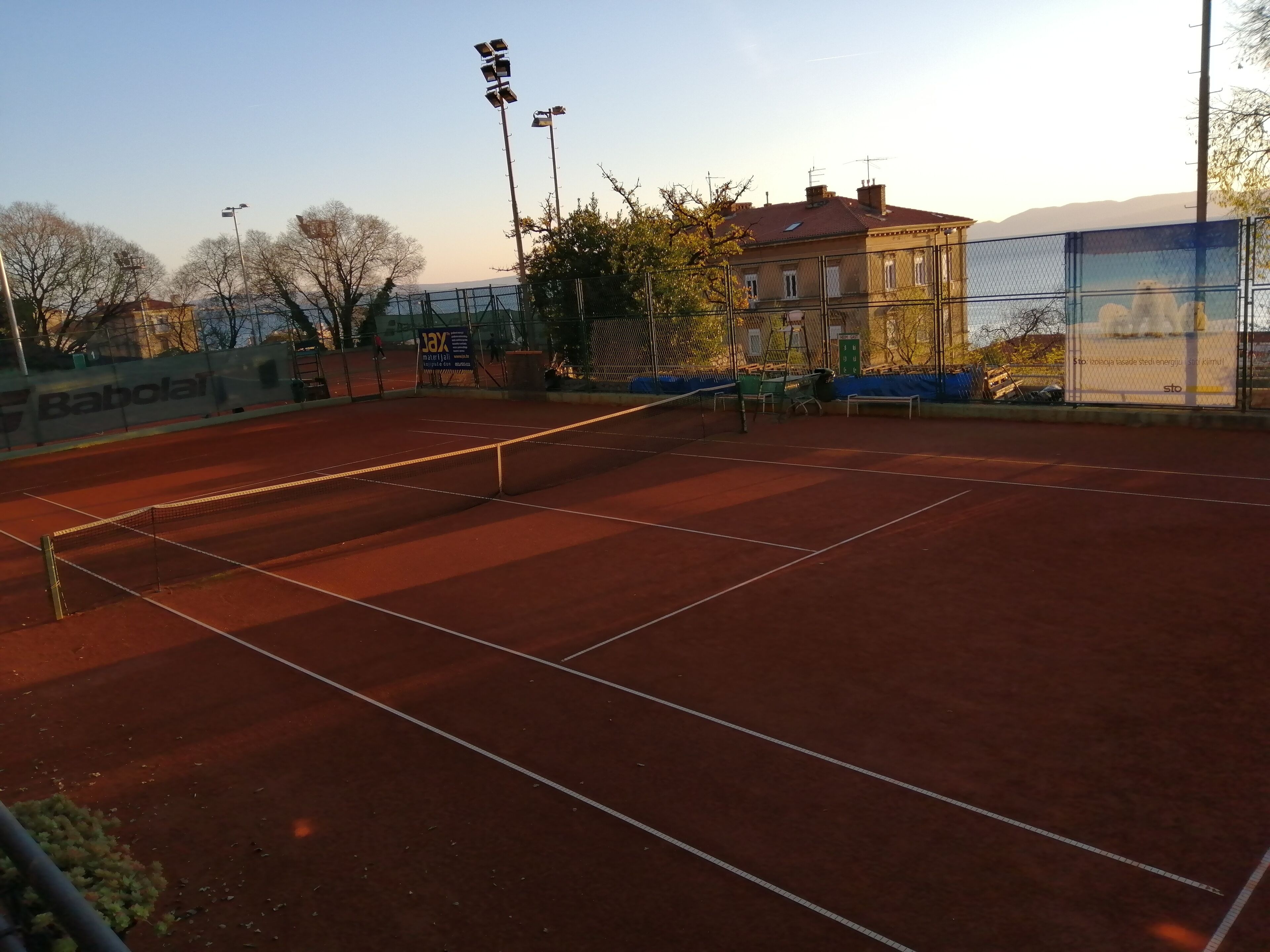 A tennis court with a net, surrounded by a fence, and a building in the background.