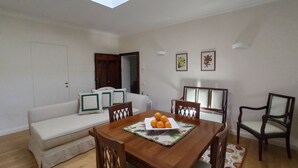 A dining area with a wooden table, chairs, and a sofa. There is a skylight above the table, and the walls are plain with a few framed pictures. - Olive Tree Suite Apartment (Piombino (LI))