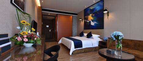 A hotel room with a bed, a desk with a vase of flowers, a chair, a round table, and a painting on the wall.