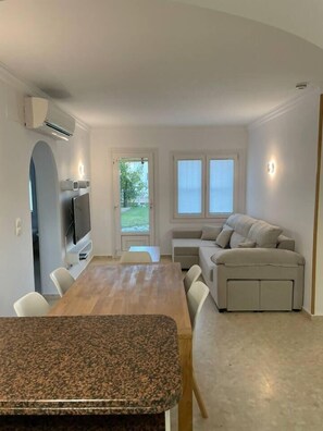 A modern living room with a wooden dining table, a beige sofa, and a TV mounted on the wall.