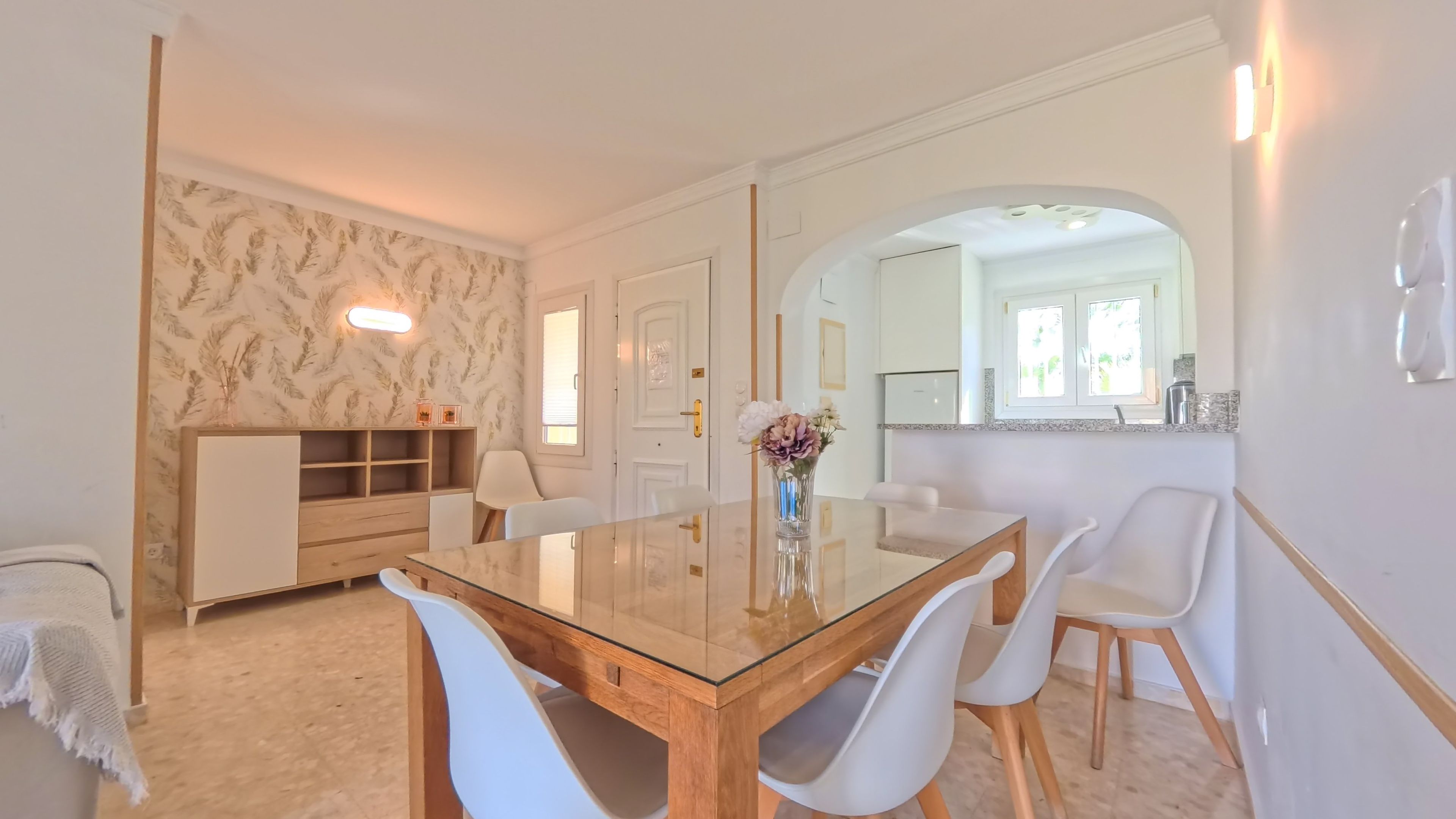 Een eetgedeelte met een tafel met glazen blad, witte stoelen, een houten kast en een bloevas.