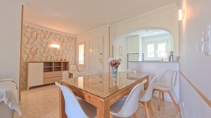 A dining area with a glass-top table, white chairs, a wooden cabinet, and a floral vase. - Brisa Azul by Midelisa. (Oliva)