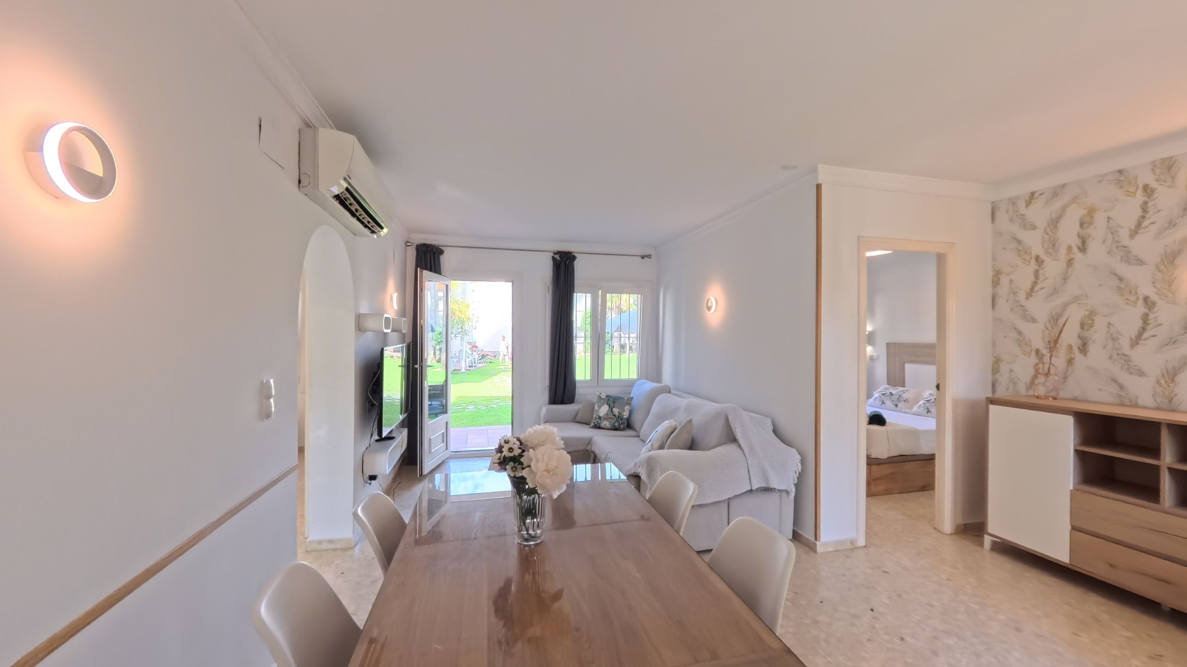 Una sala moderna con una mesa de comedor de madera, un sofá y un televisor. Hay un ventanal que ofrece vistas al exterior.