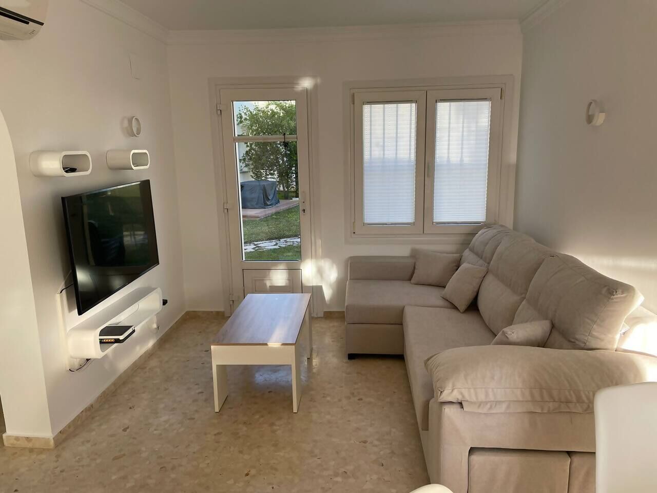 Un salón con televisor de pantalla plana, un sofá color beige, una mesa de centro y un ventanal corredero que da a un balcón.
