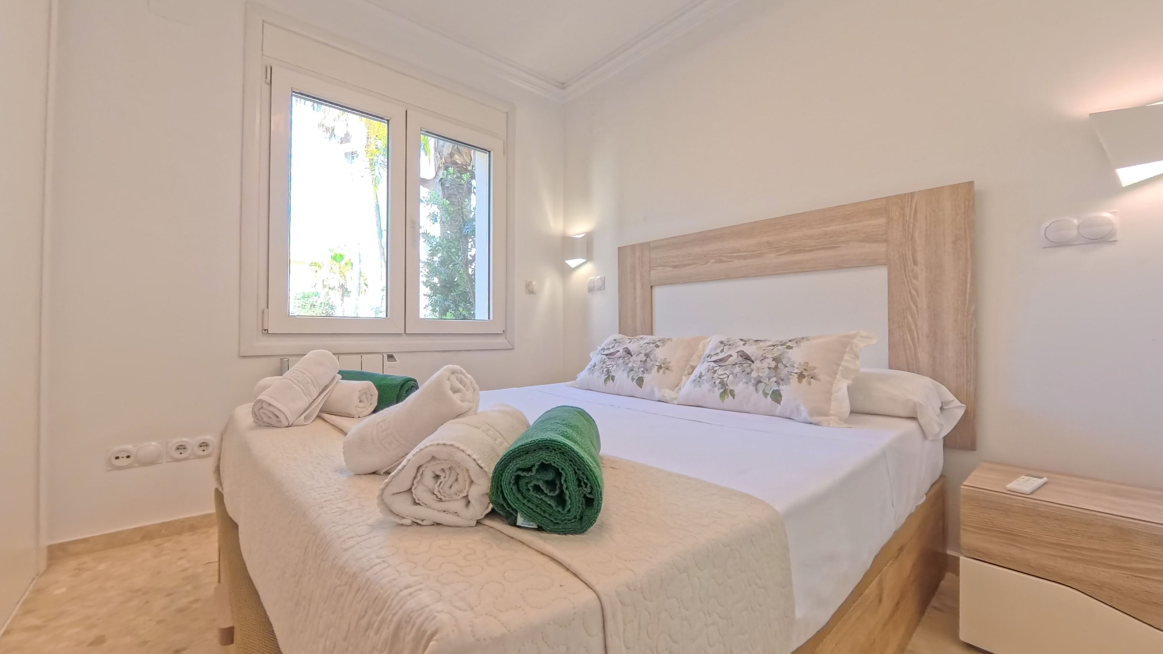 Een slaapkamer met een bed, kussens, een nachtkastje, een raam en een wandlamp.