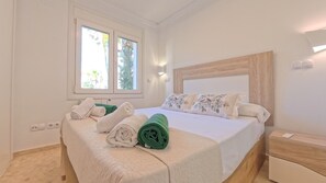 A bedroom with a bed, pillows, a nightstand, a window, and a wall-mounted light fixture. - Brisa Azul by Midelisa. (Oliva)