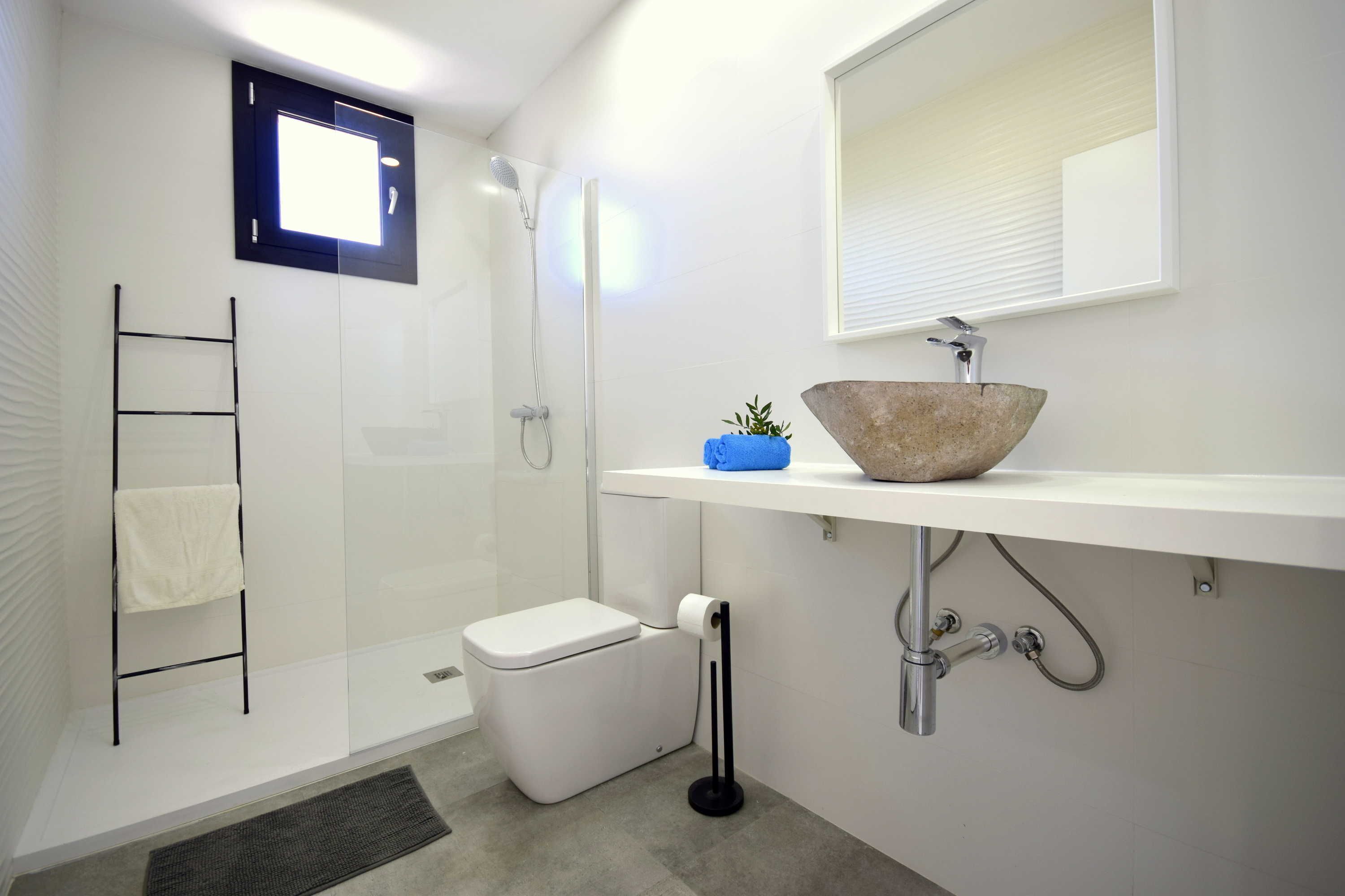 Un baño moderno con cabina de ducha de vidrio, inodoro blanco, lavabo con lavamanos de piedra y un perchero para toallas.