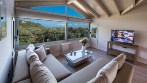 Sala de estar moderna com mesa de centro de vidro, sofá bege e televisão sobre suporte de madeira. Grandes janelas oferecem vista para uma paisagem verde.