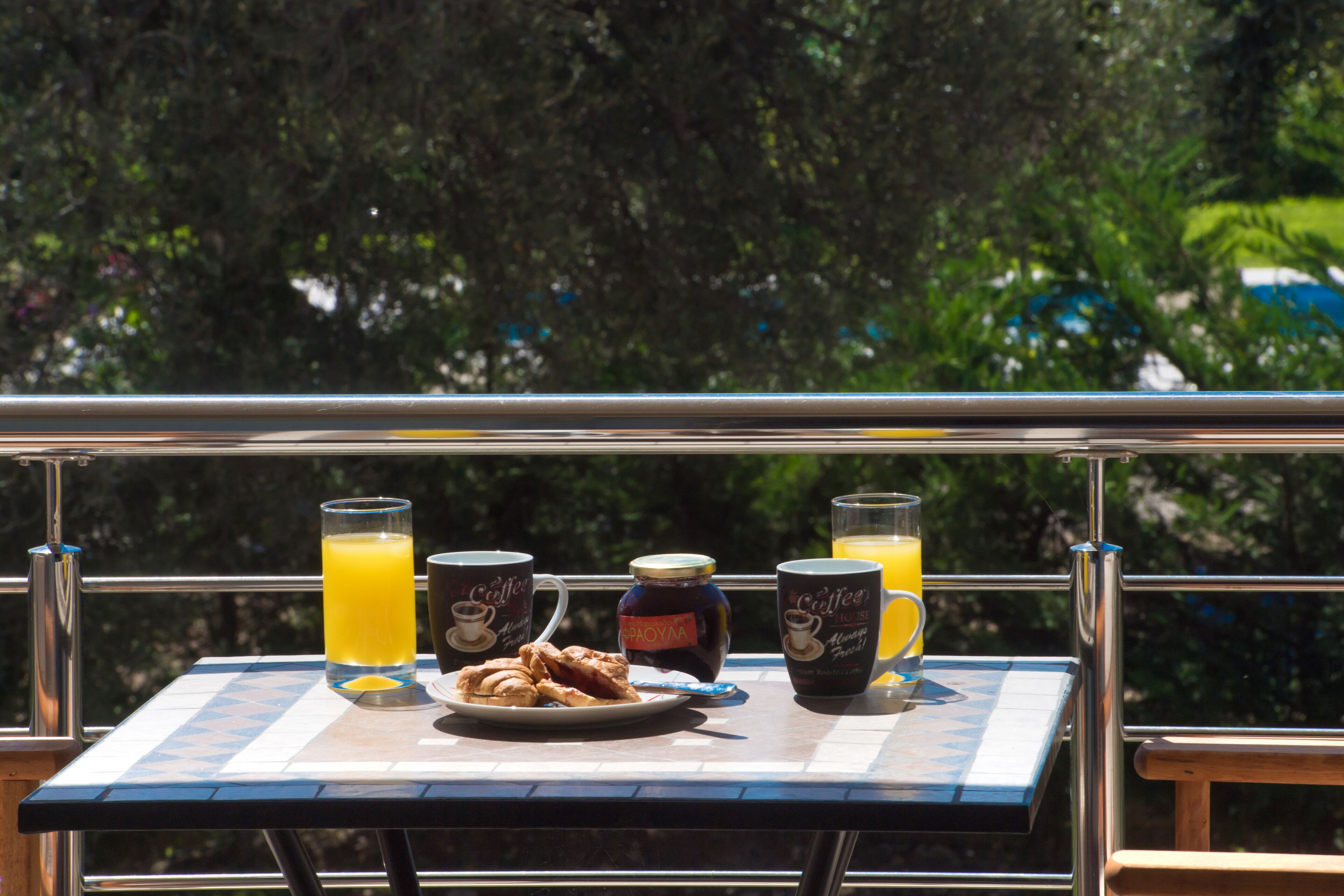 A table with breakfast items and drinks on a balcony with a metal railing and greenery in the background.