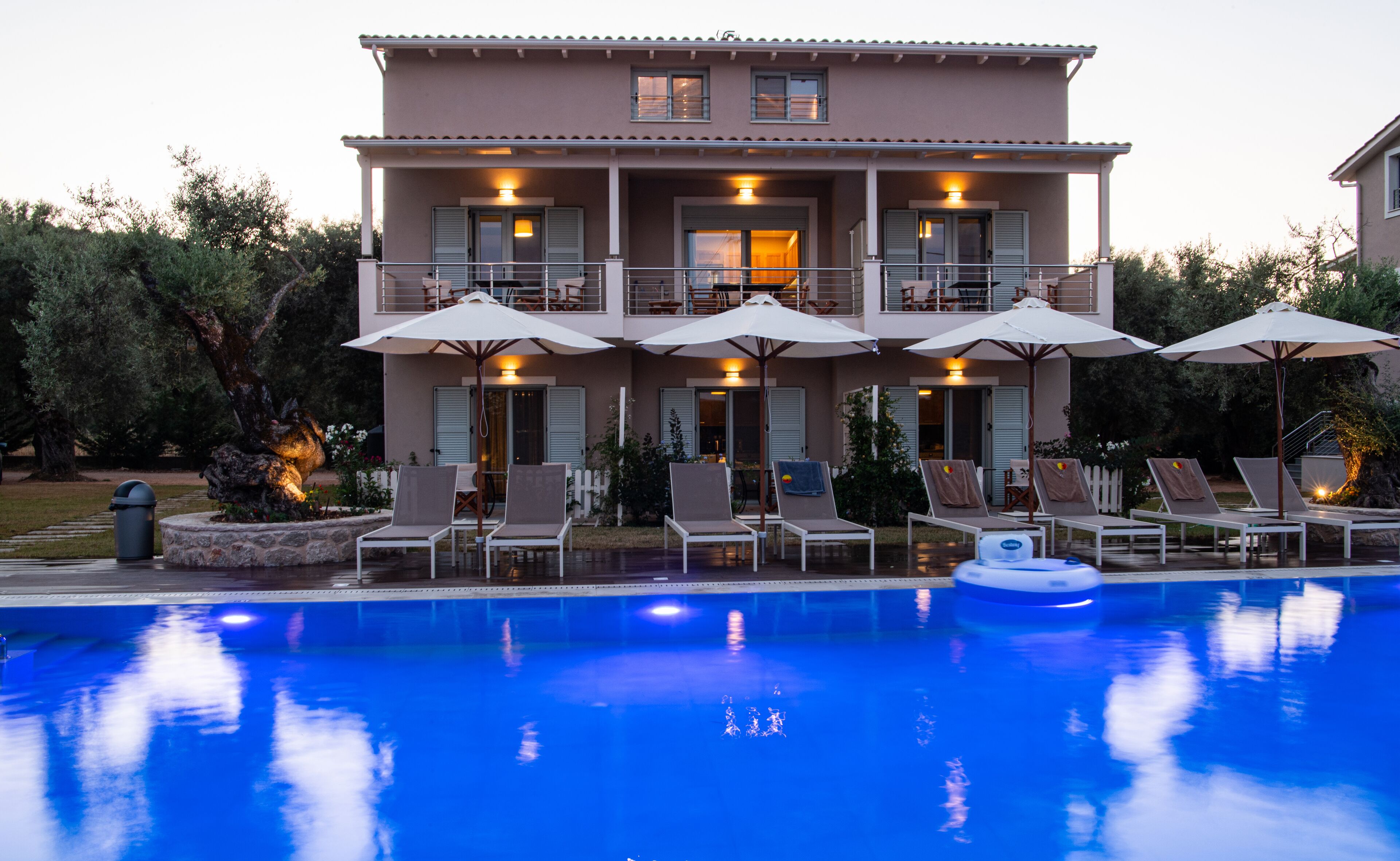Un edificio de dos pisos con piscina delantera. Hay sillones de descanso y sombrillas alrededor de la piscina.